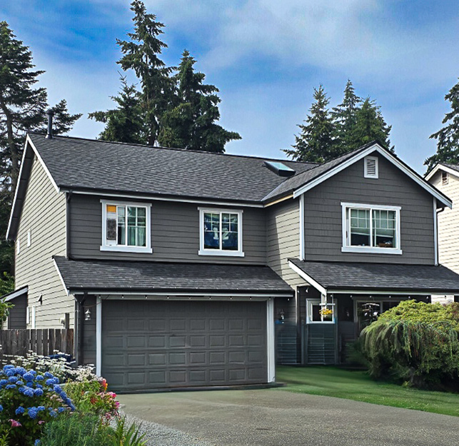 Two-story gray house with white trim, a double garage, and a driveway. Surrounded by trees, bushes, and flowers, the house has a well-maintained garden beneath a clear blue sky. Located within the Roof Boss service areas, this home exemplifies quality care from roof to curb.