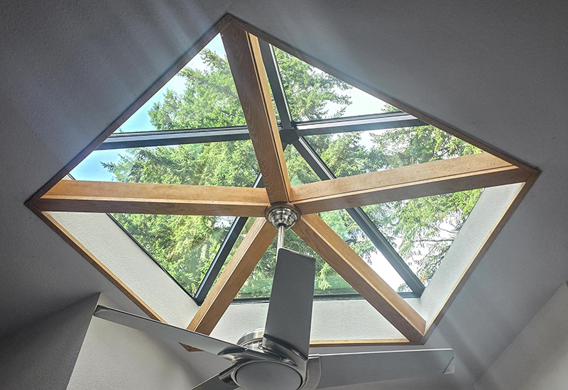 Interior view looking up through a pyramid skylight with exposed wood beams and ceiling fan