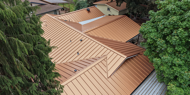 Aerial view of a house with a complex, multi-sectioned metal roof in shades of brown, surrounded by green trees and neighboring homes—perfectly showcasing the craftsmanship often seen in Roof Boss service areas.