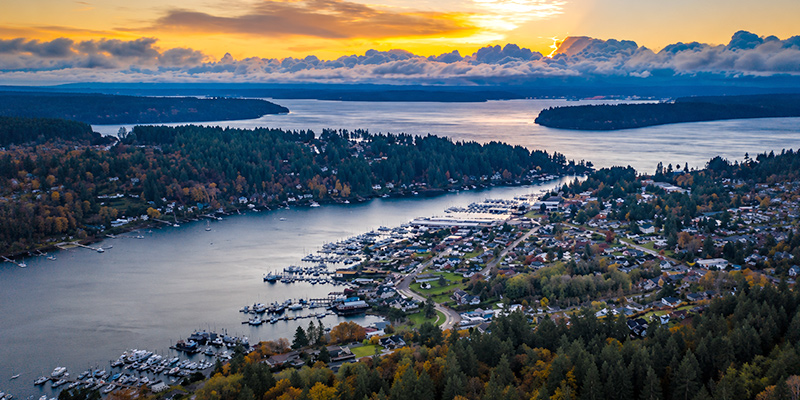 Aerial view of a coastal town with marinas, houses, and dense trees, bordered by water and forested land under a dramatic sunset sky with clouds, showcasing the picturesque landscape found within the roof boss service areas.